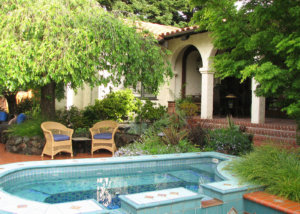 Calm rattan tea set under a shaded tree on brick open patio next to an aquamarine mosaic tile wading pool and stairs leading to shady covered back porch