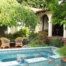 Calm rattan tea set under a shaded tree on brick open patio next to an aquamarine mosaic tile wading pool and stairs leading to shady covered back porch