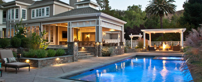 Luxurious backyard featuring a sparkling rectangular pool surrounded by a stone patio with elegant outdoor lighting. The space includes a covered outdoor kitchen and bar area attached to a beautiful house with large windows and classic architecture. To the right, a cozy pergola with seating and a fire pit creates an inviting lounge area. The landscape is enhanced with lush greenery, ornamental grasses, and a mix of shrubs, blending the outdoor living space seamlessly with the surrounding natural environment.