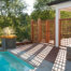 Poolside deck area featuring wooden lattice privacy screens casting intricate shadows on the stone patio. A tall modern planter with a spiky plant adds contrast, while the crystal-clear pool reflects the serene, tree-filled backdrop