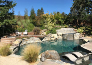 Open gray stone tile patio with curvy stone lined pool and raised jacuzzi with lounge chairs on staged wooden deck