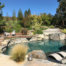 Open gray stone tile patio with curvy stone lined pool and raised jacuzzi with lounge chairs on staged wooden deck