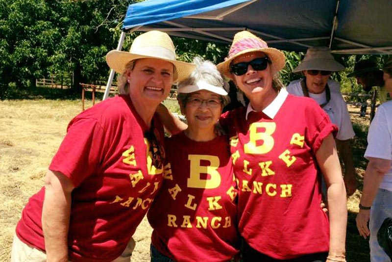 Three women smiling and posing together under a canopy at an outdoor event. They are wearing matching red shirts with a large yellow 'B' and the words 'Walker Ranch' printed on them. Each woman is wearing a sun hat, and there are trees and other people in the background at a non profit eventfor Adults with Autism – harvesting a sense of purpose.