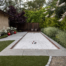 A beautifully landscaped backyard featuring a pristine bocce ball court lined with a smooth gravel playing surface, surrounded by a clean concrete border. The court is tucked between lush green shrubs and ornamental grasses that add texture and color to the space. A row of delicate white flowers lines one side, planted against the tan wall of a nearby structure. Tall trees, including a deep purple-leafed tree, provide shade and a sense of privacy, while bright red planters punctuate the landscape with pops of color. At the far end of the court, a pair of wooden chairs invites relaxation and socializing in this elegant, tranquil setting.