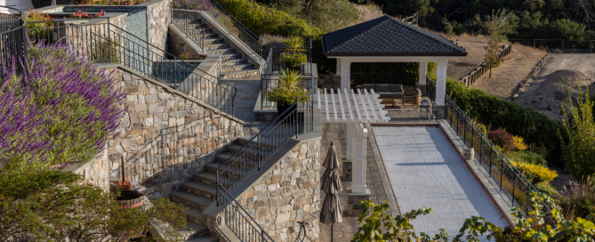 Elegant multi-level backyard terrace featuring stone retaining walls, metal railings, and winding staircases that lead to various outdoor areas. The space includes a bocce ball court with a white surface, flanked by a charming pavilion with a dark roof, offering a shaded spot for relaxation. Lush landscaping with colorful flowers, greenery, and strategically placed planters adds vibrancy to the stonework. The elevated design provides stunning views of the surrounding hills and natural landscape, creating a luxurious and inviting outdoor retreat.