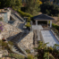 Elegant multi-level backyard terrace featuring stone retaining walls, metal railings, and winding staircases that lead to various outdoor areas. The space includes a bocce ball court with a white surface, flanked by a charming pavilion with a dark roof, offering a shaded spot for relaxation. Lush landscaping with colorful flowers, greenery, and strategically placed planters adds vibrancy to the stonework. The elevated design provides stunning views of the surrounding hills and natural landscape, creating a luxurious and inviting outdoor retreat.