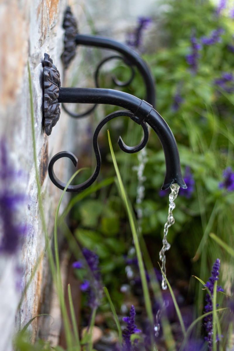 A close-up of a decorative wall fountain featuring elegant black iron spouts with intricate curved designs. Water gently flows from the spouts into a garden bed filled with vibrant purple flowers and lush green foliage. The fountain is set against a natural stone wall, adding texture and rustic charm to the scene. The combination of flowing water, ornamental ironwork, and blooming plants creates a peaceful and serene garden ambiance, perfect for relaxation and adding a touch of elegance to the outdoor space.