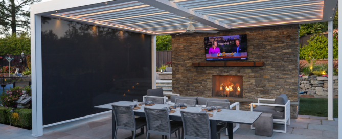 A modern outdoor entertainment space features a sleek, white pergola with retractable overhead slats for adjustable lighting. Beneath the pergola is a stylish dining area, complete with a long, rectangular table and six contemporary gray chairs. Adjacent to the dining area is a cozy lounge space with cushioned seating, all facing a stone fireplace with a warm, glowing fire. Above the fireplace, a large television is mounted, creating a perfect spot for watching shows or sports outdoors. The space is bordered by lush landscaping and stonework, adding a touch of natural elegance.