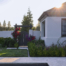 A serene and elegant backyard garden featuring a modern home with a light-colored exterior and large black-framed windows. The garden includes a well-manicured lawn with stepping stone paths leading to a dark wooden gate framed by blooming bougainvillea, creating a beautiful entryway. Lush green hedges and colorful flowering plants line the edges of the lawn, adding vibrant texture and color to the space. In the foreground, a sleek patio and pool provide a luxurious outdoor living area, while the soft sunlight filtering through the trees enhances the peaceful ambiance of the scene.