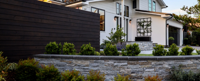 A beautifully landscaped modern home with a clean white exterior and large black-framed windows. The front yard features a low stone retaining wall with neatly trimmed shrubs and vibrant green plants, creating a structured yet natural look. A dark wooden privacy fence adds contrast to the home's light facade while providing seclusion. The house's architecture is contemporary with sharp lines and large windows that allow plenty of natural light to flood the interior. The surrounding trees and well-maintained garden beds add warmth and softness to the modern, sleek design of the property.