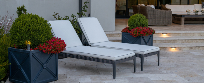 A sophisticated outdoor patio featuring two cushioned lounge chairs with white upholstery, perfect for relaxing. The chairs are flanked by stylish dark blue planters filled with neatly trimmed boxwood shrubs and vibrant red flowers, adding a pop of color to the neutral-toned space. The patio is paved with light stone tiles, leading to a set of steps that connect to a spacious seating area in the background. The large glass doors of the house reflect the warm light from the interior, creating a cozy and inviting ambiance. The overall design combines elegance and comfort, ideal for outdoor leisure and entertaining.