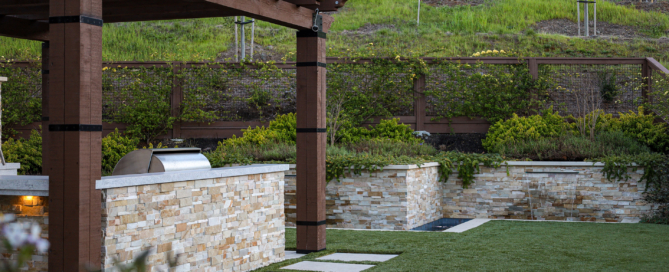 Outdoor kitchen with stone bar, pergola, and built-in water feature by Montgomery Robbins – modern backyard landscape design