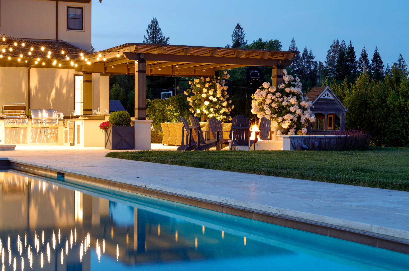 Elegant backyard design by Montgomery Robbins with a pergola-covered outdoor kitchen, fire pit seating, and poolside ambiance at dusk.