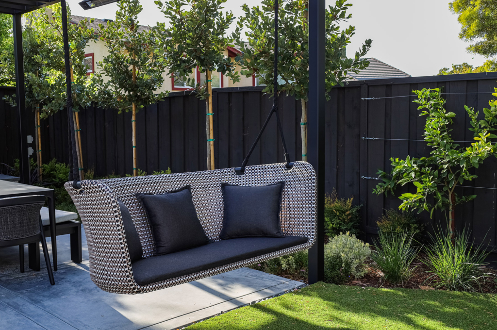 Modern outdoor hanging loveseat with a woven design, featuring two plush black cushions, suspended from a sturdy metal frame. The swing overlooks a well-maintained lawn and sits beneath partial shade, offering a cozy seating area. The surrounding black wooden fence adds contrast, while young trees and lush greenery enhance the tranquil backyard atmosphere.