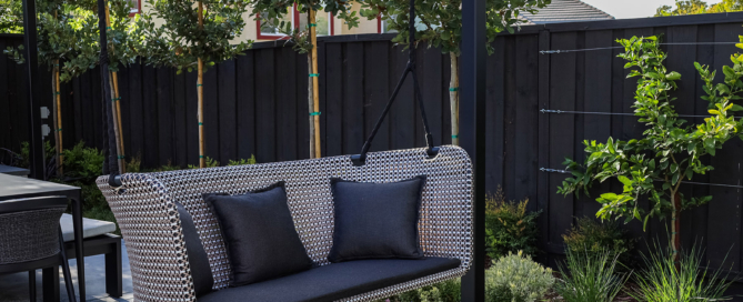 Modern outdoor hanging loveseat with a woven design, featuring two plush black cushions, suspended from a sturdy metal frame. The swing overlooks a well-maintained lawn and sits beneath partial shade, offering a cozy seating area. The surrounding black wooden fence adds contrast, while young trees and lush greenery enhance the tranquil backyard atmosphere.