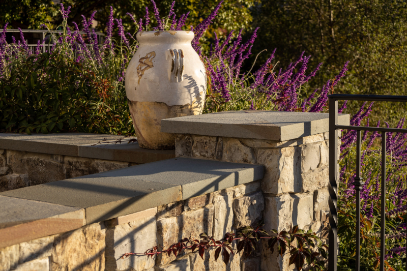 A tranquil outdoor scene featuring a rustic stone wall topped with smooth stone caps, bathed in warm sunlight. A weathered ceramic urn with artistic markings sits atop the wall, adding an earthy and artistic touch to the setting. Behind the wall, vibrant purple flowers create a striking contrast with the natural stone, adding color and life to the space. A black metal railing runs alongside the stone structure, blending with the overall rustic yet elegant garden aesthetic. The soft lighting and shadow play create a peaceful, inviting atmosphere in this beautifully landscaped area.