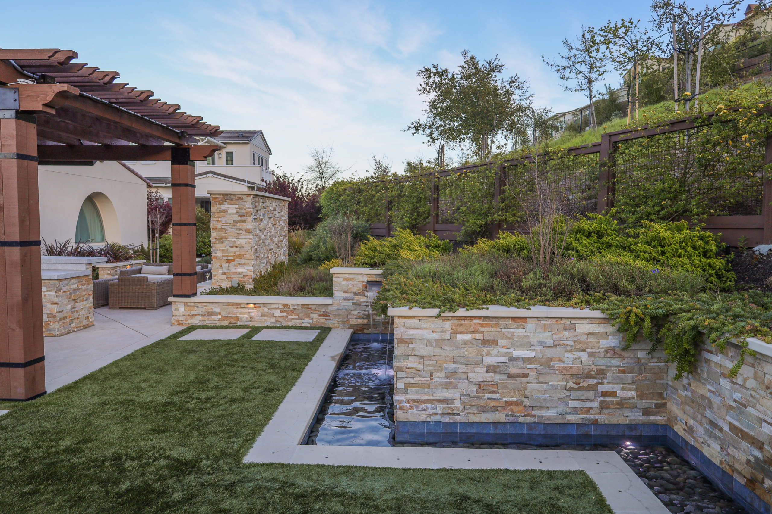 Montgomery Robbins, Inc. landscape architecture design in Northern California showcasing a modern backyard with stone retaining walls, integrated water feature, wood pergola, artificial turf, and layered hillside plantings for a luxury outdoor living space
