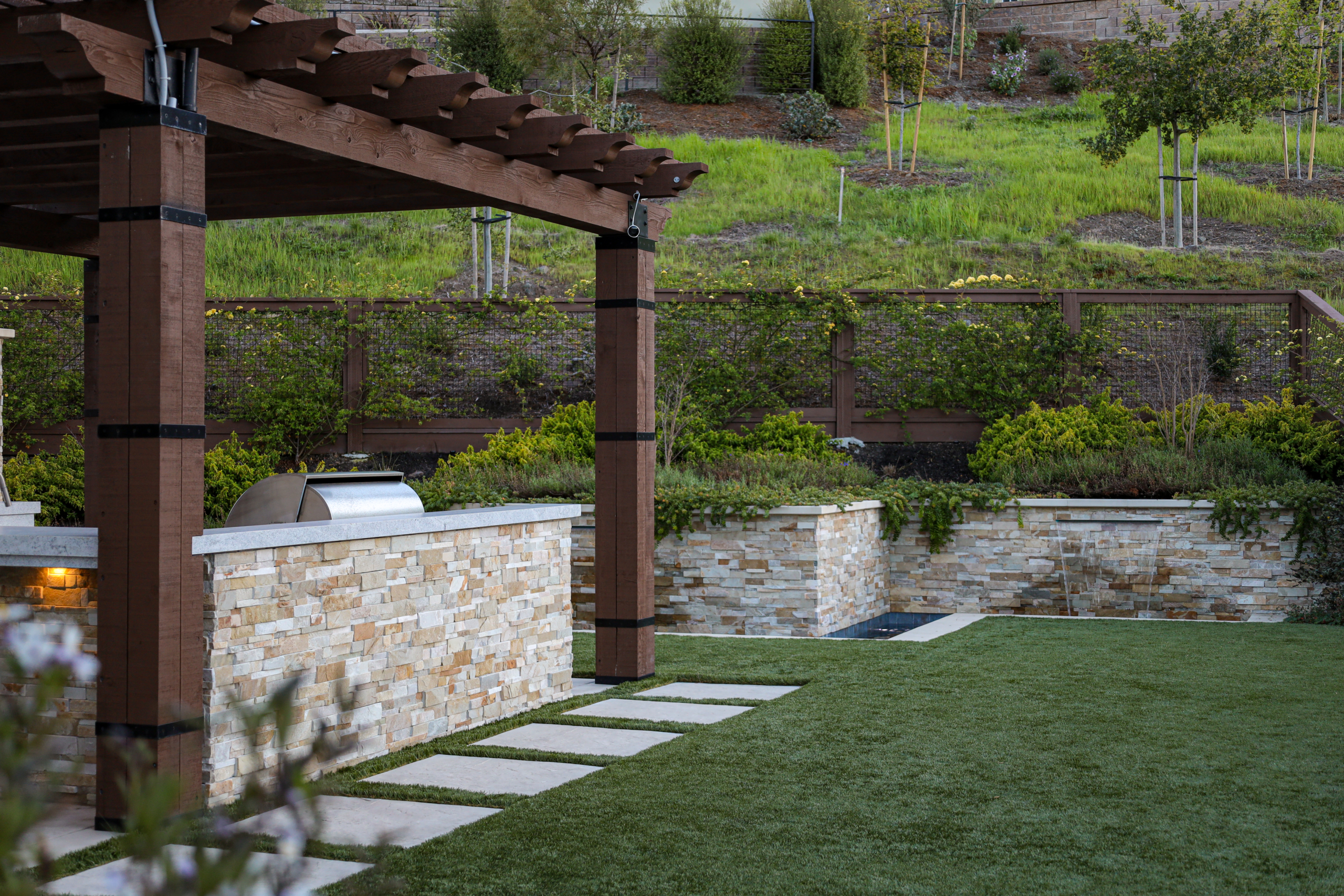 Montgomery Robbins, Inc. landscape architecture design featuring a custom outdoor kitchen with natural stone veneer, wood pergola, built-in stainless steel grill, modern stepping stone pathway, and lush hillside planting in a luxury Bay Area backyard.
