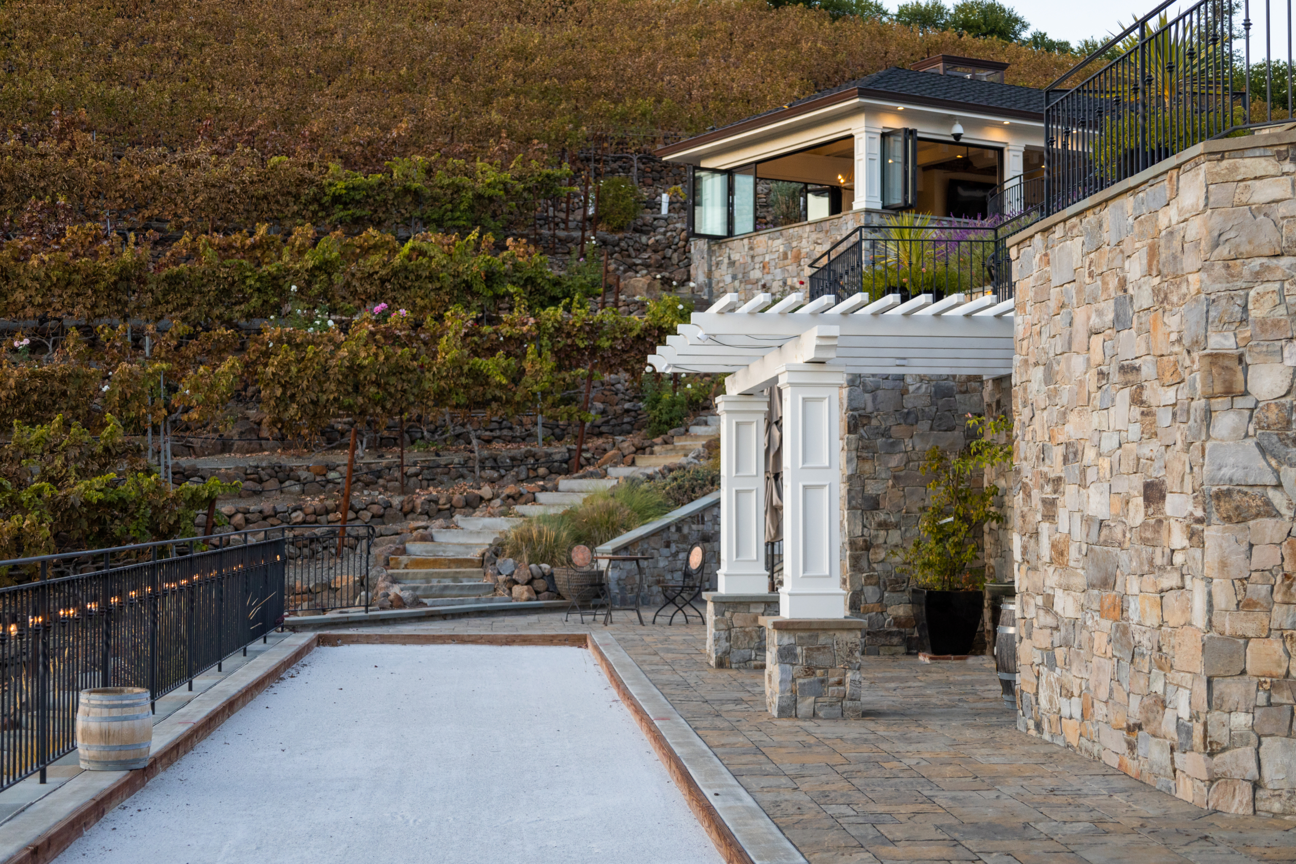 Hillside vineyard landscape in Northern California designed by Montgomery Robbins, Inc., featuring a terraced stone retaining wall, elegant white pergola, and bocce court overlooking a luxury outdoor pavilion. This Bay Area residential landscape architecture project blends natural stonework with formal design elements to create a refined yet relaxed “California Casual” setting that celebrates the region’s vineyard topography and outdoor living lifestyle.