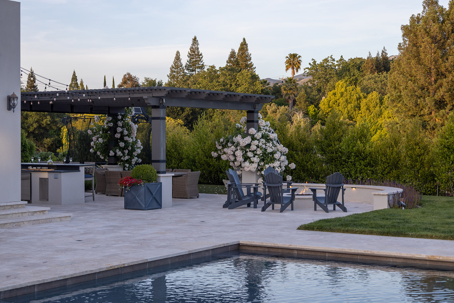 Outdoor patio area with a luxurious pool in the foreground, featuring a pergola-covered seating area adorned with string lights and white flowering plants. The patio is furnished with gray Adirondack chairs surrounding a small fire pit and a dining area under the pergola. The lush green trees and hilly landscape in the background provide a scenic, tranquil setting.