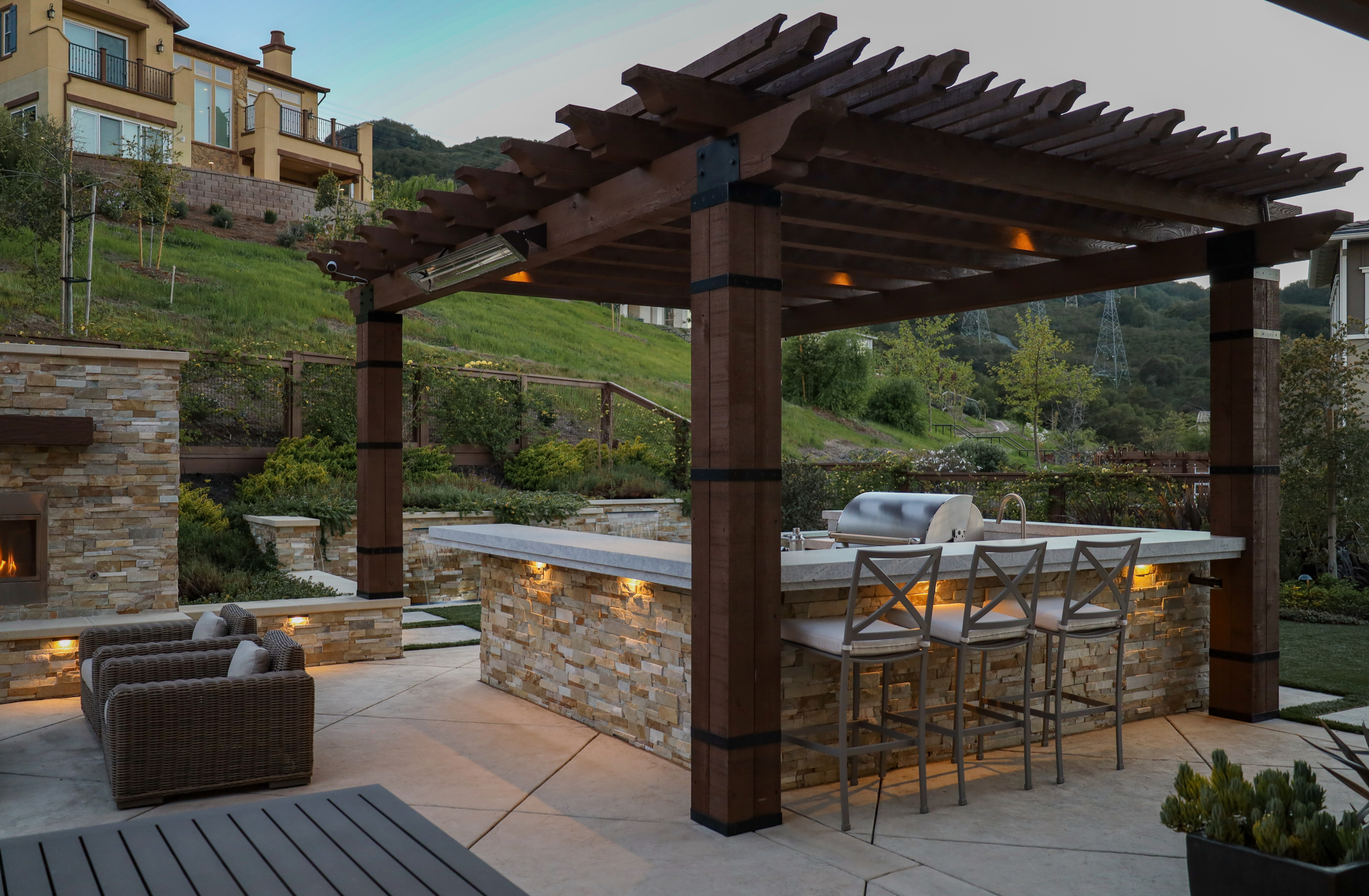 Outdoor kitchen and bar area under wood pergola by Montgomery Robbins, featuring stone accents and modern landscaping.
