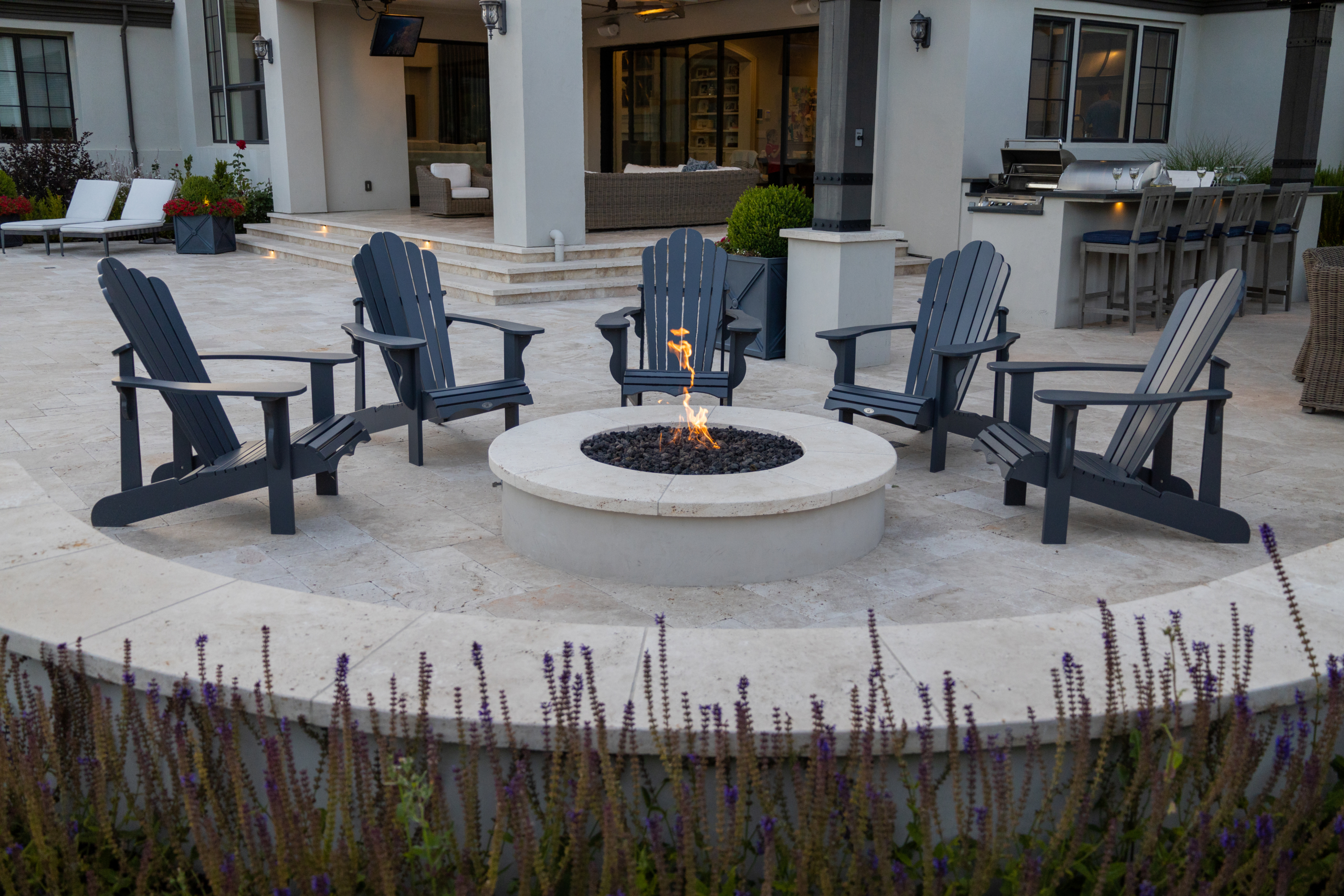 Modern fire pit seating area with Adirondack chairs by Montgomery Robbins, set in a luxury outdoor patio with kitchen and lounge.
