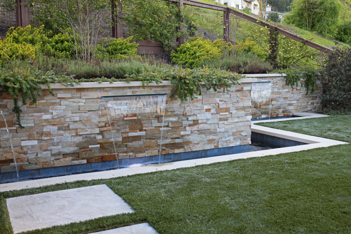Contemporary water feature with tiered stone walls and cascading fountains by Montgomery Robbins – modern backyard landscape design with lush greenery