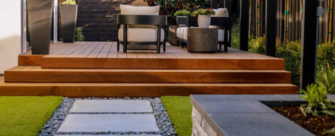 Elegant wood deck with full-length steps meets with contemporary concrete pavers, faux turf and river rock detailing