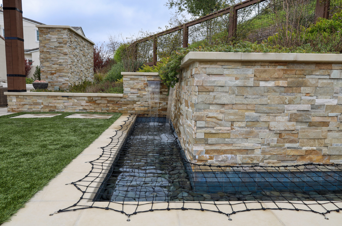 Modern water feature with stacked stone walls and safety netting by Montgomery Robbins – custom backyard landscape design with built-in fountain