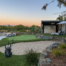 Backyard with a luxurious golf-themed setup featuring a putting green and a sand bunker, surrounded by manicured landscaping and stone accents. In the foreground, a golf bag with clubs sits on the green. The background includes a modern outdoor entertainment space with a large screen under a shaded pergola, accompanied by lounge chairs for relaxation. The entire area is framed by natural greenery, creating a serene and sophisticated environment, perfect for golf practice and outdoor leisure. The sunset sky adds a warm, inviting glow to the scene.