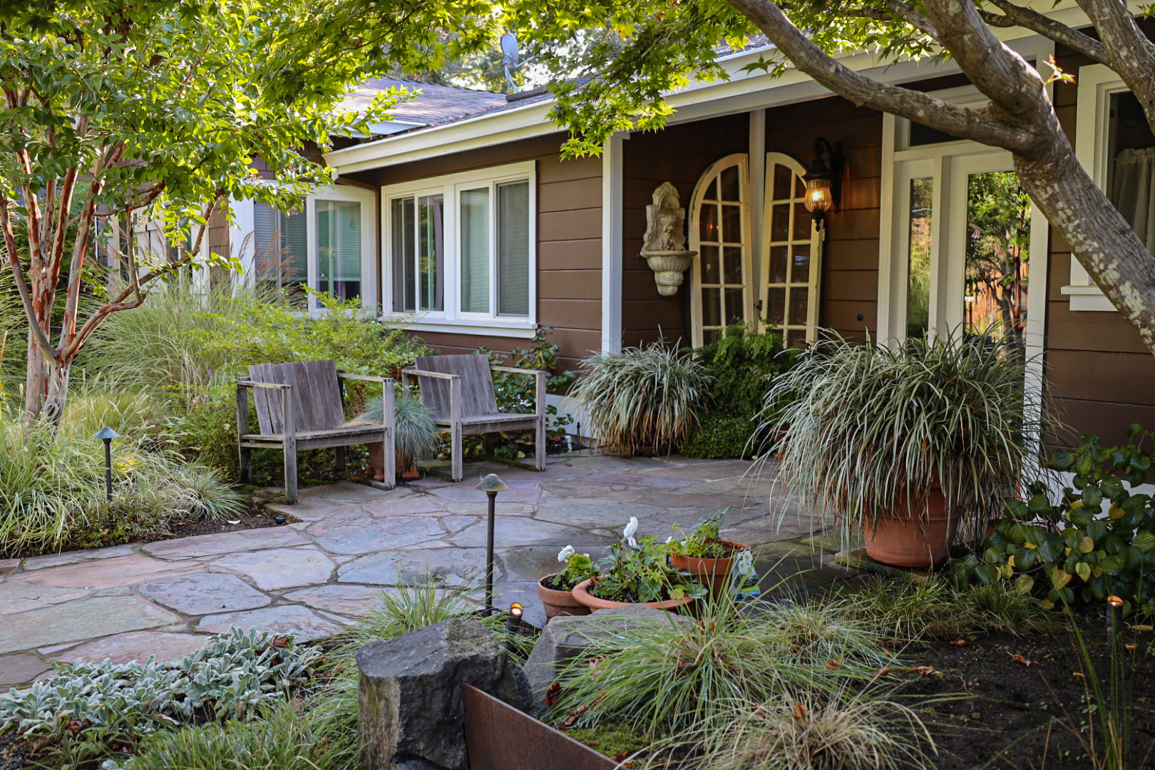 Shaded garden entryway by Montgomery Robbins featuring flagstone paving, rustic seating, ornamental grasses, and mature trees for a peaceful front yard retreat.