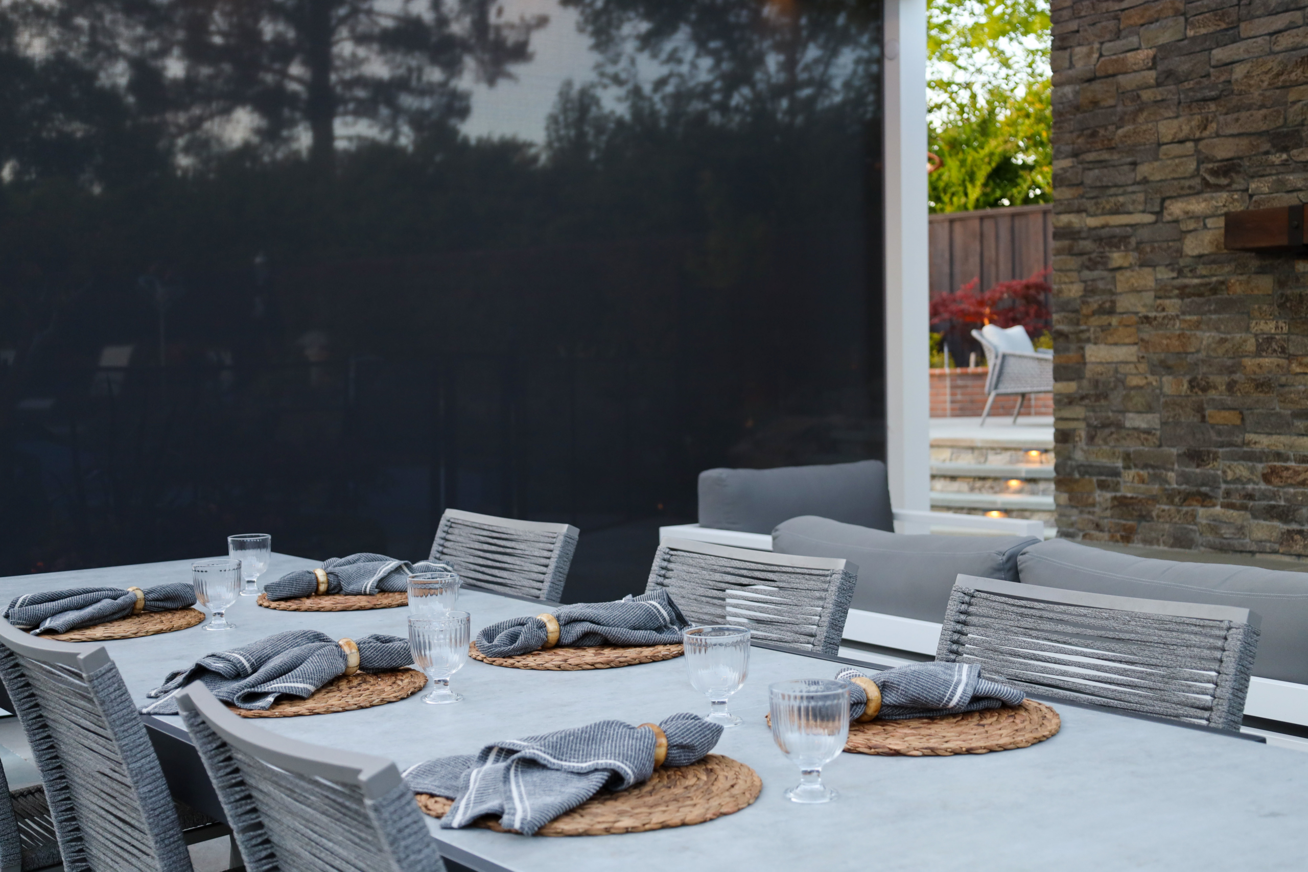 Elegant outdoor dining table with woven placemats and gray seating set against a stone fireplace by Montgomery Robbins
