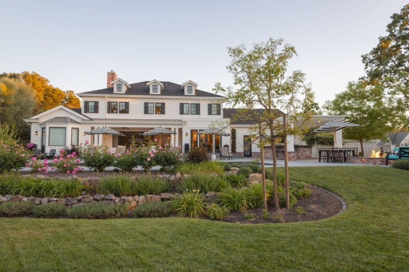 Spacious backyard of a stunning two-story farmhouse-style home with a well-manicured lawn and lush garden beds filled with vibrant flowers, ornamental grasses, and small trees. The house features white siding, black shutters, and dormer windows, creating a classic and elegant look. An outdoor seating area, fire pit, and pergola can be seen on the right, enhancing the property's inviting and family-friendly atmosphere. Mature trees and carefully landscaped spaces add to the serene and picturesque setting.