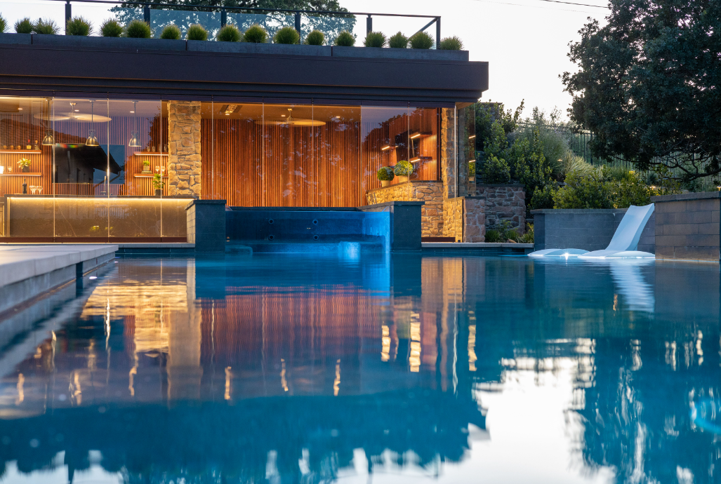 Modern luxury swimming pool with waterfall feature reflecting a warm, illuminated outdoor pavilion, designed by Montgomery Robbins, Inc. in the Bay Area. The sleek hardscape, seamless indoor-outdoor connection, and refined planting design showcase high-end residential landscape architecture tailored to Northern California living.