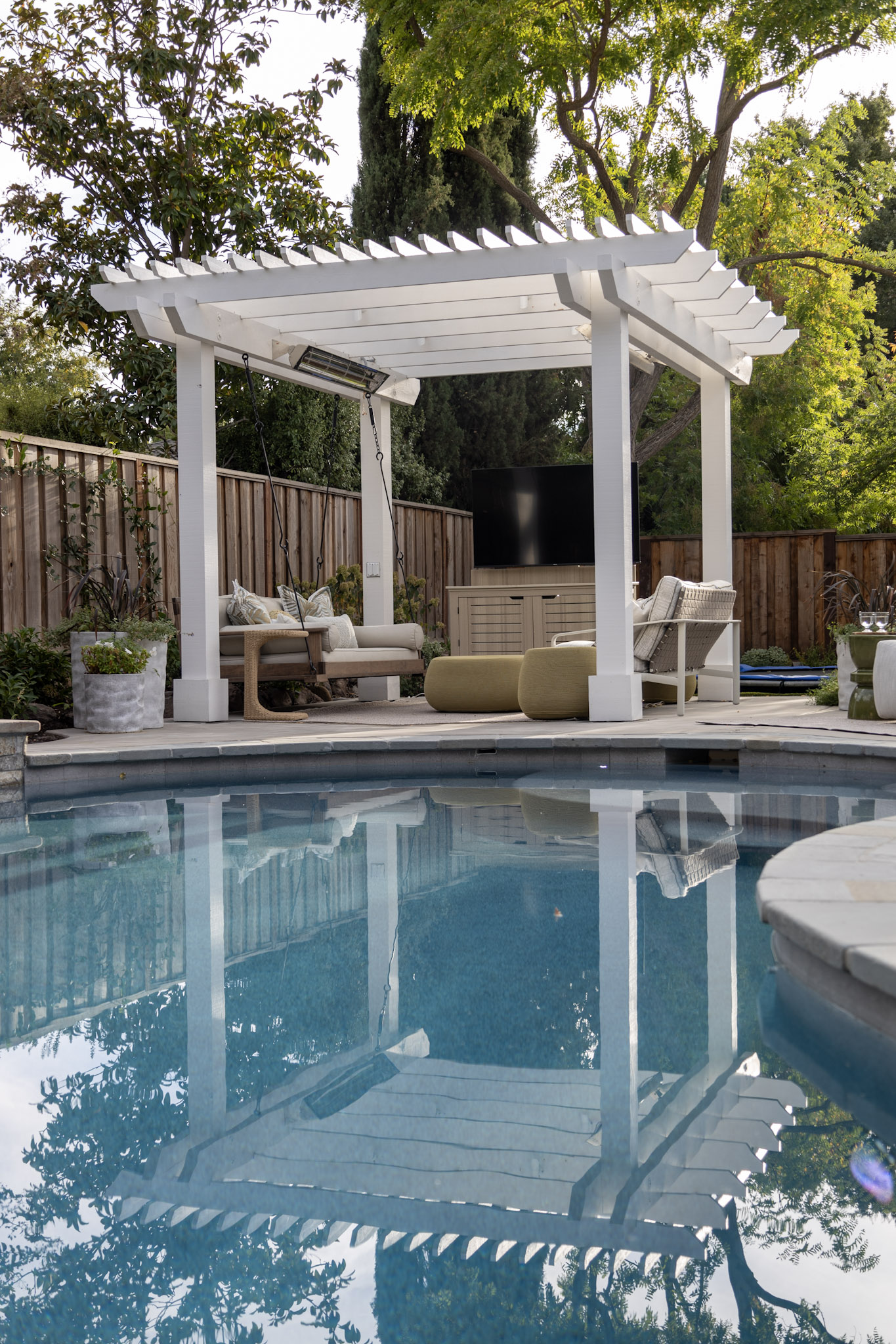 Modern backyard pool area featuring a white pergola with a cozy seating arrangement, including a sofa with patterned cushions, a lounge chair, and green ottomans. The pergola provides shade over the seating space, which faces an outdoor TV mounted on a cabinet. Lush greenery and tall trees surround the area, with a wooden fence in the background, creating a serene and private atmosphere. The clear pool in the foreground reflects the pergola and the surrounding foliage, enhancing the tranquil and inviting ambiance.