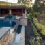 The image showcases a luxurious backyard featuring a modern pool with a sleek, angular design and integrated spa. The pool is surrounded by elegant stonework and raised planters, adding texture and visual interest. A contemporary pool house with glass walls, stone accents, and wood paneling overlooks the water, offering an inviting space for relaxation and entertainment. The landscape includes neatly arranged ornamental grasses and shrubs, creating a natural transition from the manicured pool area to the hillside beyond. Mature trees and soft evening light enhance the tranquil ambiance of this sophisticated outdoor retreat.