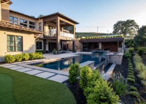 The image showcases a stunning backyard of a modern luxury home with a beautiful pool and spa. The pool features a sleek, linear design with clean edges and integrated stone accents, creating a sophisticated look. Adjacent to the pool is a well-manicured garden with lush green shrubs, ornamental grasses, and decorative lighting, enhancing the serene atmosphere. The home’s exterior boasts large windows, a spacious balcony, and a covered patio area with stylish seating, perfect for outdoor living. In the background, a contemporary pool house with wood and stone elements provides additional entertainment space, seamlessly blending with the landscape.