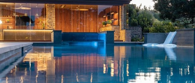 The image depicts a luxurious, modern backyard with a stylish outdoor living space featuring a pool in the foreground. The centerpiece is a sleek pool house with floor-to-ceiling glass walls, stone accents, and rich wooden paneling. Inside, warm lighting highlights elegant decor with hanging pendant lights and shelves adorned with plants and glassware. A modern fire feature adds a sophisticated touch, creating a cozy atmosphere. The rooftop garden is lined with lush greenery, enhancing the serene and upscale design.