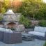 Outdoor living area featuring a custom stone fireplace with built-in seating ledges, surrounded by tiered planting beds, ornamental trees, and a flagstone patio with modern woven furniture; showcases thoughtful landscape architecture with structured hardscaping, layered greenery, and intimate entertainment design.