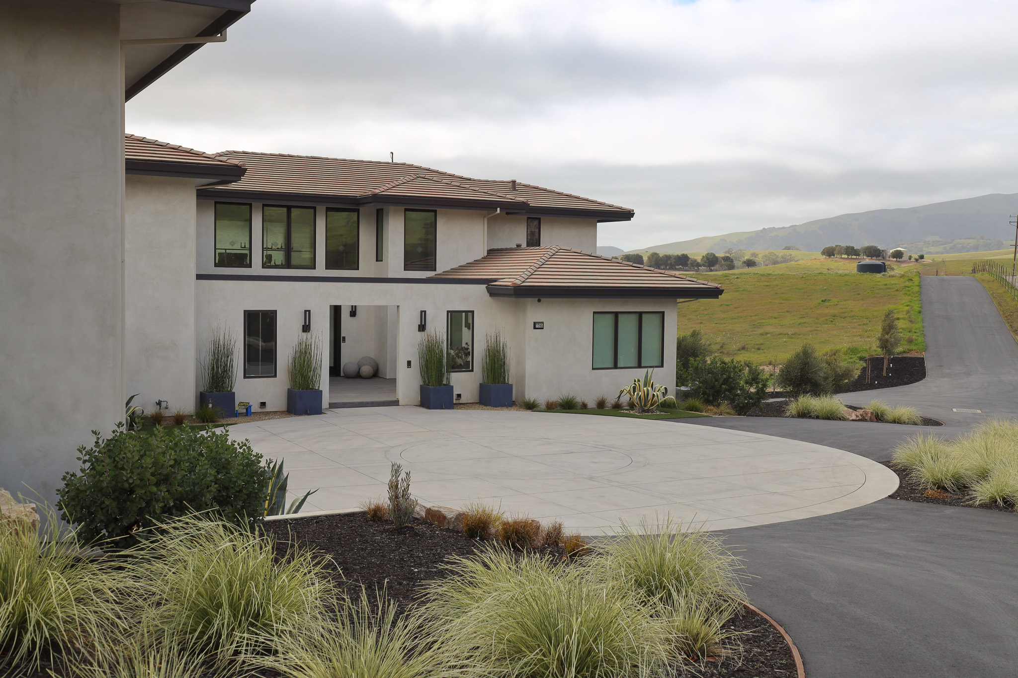 Modern front yard with firescaping landscape design featuring drought-tolerant, fire-resistant plants such as ornamental grasses and succulents, wide concrete driveway acting as a defensible space, non-combustible mulch, and strategic plant placement away from the home’s structure to enhance wildfire resilience.