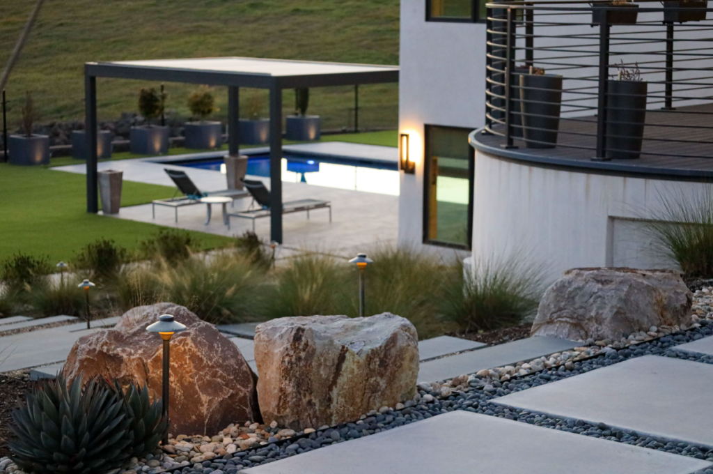 Modern outdoor pool and pavilion with drought-tolerant landscaping — Agave, ornamental grasses, and low groundcover planted in mulched beds along a stone retaining wall, reflecting drought-tolerant plant choices suited to Northern California's Sunset Zone 14–17 / USDA Zone 9b–10a wine country climate.