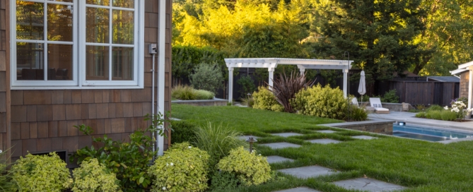 Custom residential landscape architecture by Montgomery Robbins, Inc. featuring a modern stepping-stone pathway set in lush green lawn, layered planting beds with shrubs and ornamental grasses, and a view toward a luxury backyard pool and pergola lounge area framed by privacy trees for timeless Northern California outdoor living.