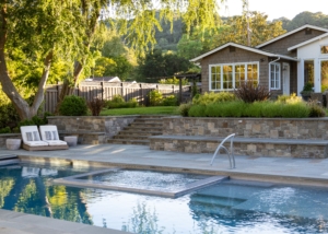 Modern backyard pool and garden design by Montgomery Robbins, Inc., a leading residential landscape architecture firm in Alamo, California. This luxury outdoor living space features a sleek swimming pool with integrated spa, custom stone hardscape, terraced retaining walls, and layered plantings that frame the home. Comfortable lounge seating, lush greenery, and seamless transitions from poolside to elevated garden areas showcase Montgomery Robbins’ expertise in creating high-end residential landscapes that balance beauty, function, and California lifestyle design.