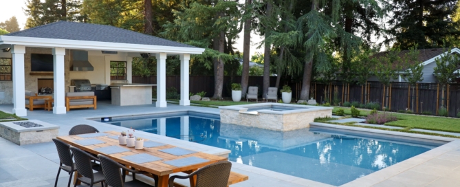 Luxury residential backyard featuring a modern swimming pool with integrated spa, outdoor kitchen pavilion, and teak dining space, designed with premium hardscape materials and refined spatial planning. Surrounded by mature trees and layered plantings, the space reflects high-end landscape architecture principles with clean lines, balanced proportions, and seamless indoor-outdoor living. Invisible, high-performance drainage solutions are integrated into the stone paving and lawn areas to manage stormwater discreetly while maintaining a polished aesthetic and long-term functionality for Northern California residential environments.